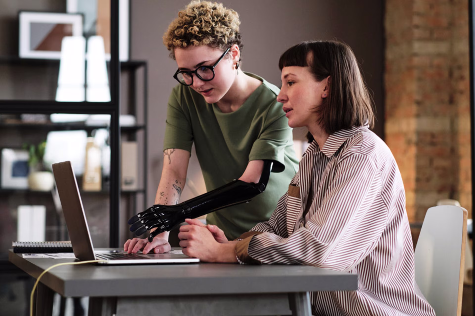 a11y Young woman with prosthetic arm pointing at laptop and talking to her colleague during work at office